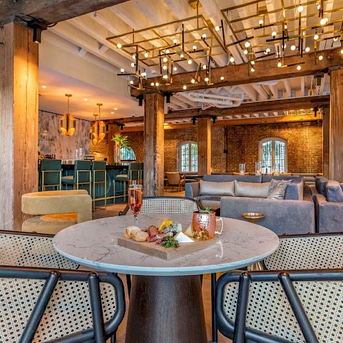 The image shows a stylish restaurant or lounge with a modern interior design, featuring a table with chairs, cozy seating areas, and a well-equipped bar.