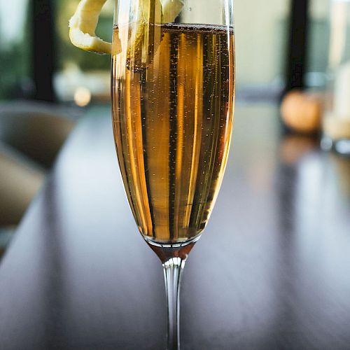 A tall champagne flute filled with a bubbly drink, garnished with a lemon twist, sits on a dark surface in a modern setting.