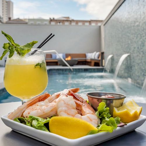 The image shows a refreshing drink and a plate of shrimp with lemon wedges on a table next to a pool with waterfalls.