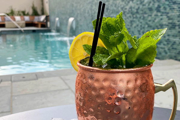 A copper mug with a drink, garnished with mint and lemon, sits by a poolside with lounge chairs and water features in the background.
