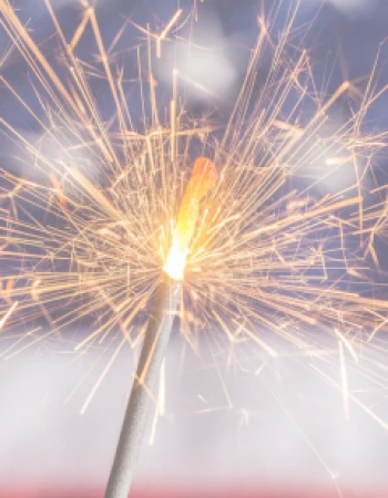 A lit sparkler is captured in the foreground, with an American flag pattern blurred in the background.