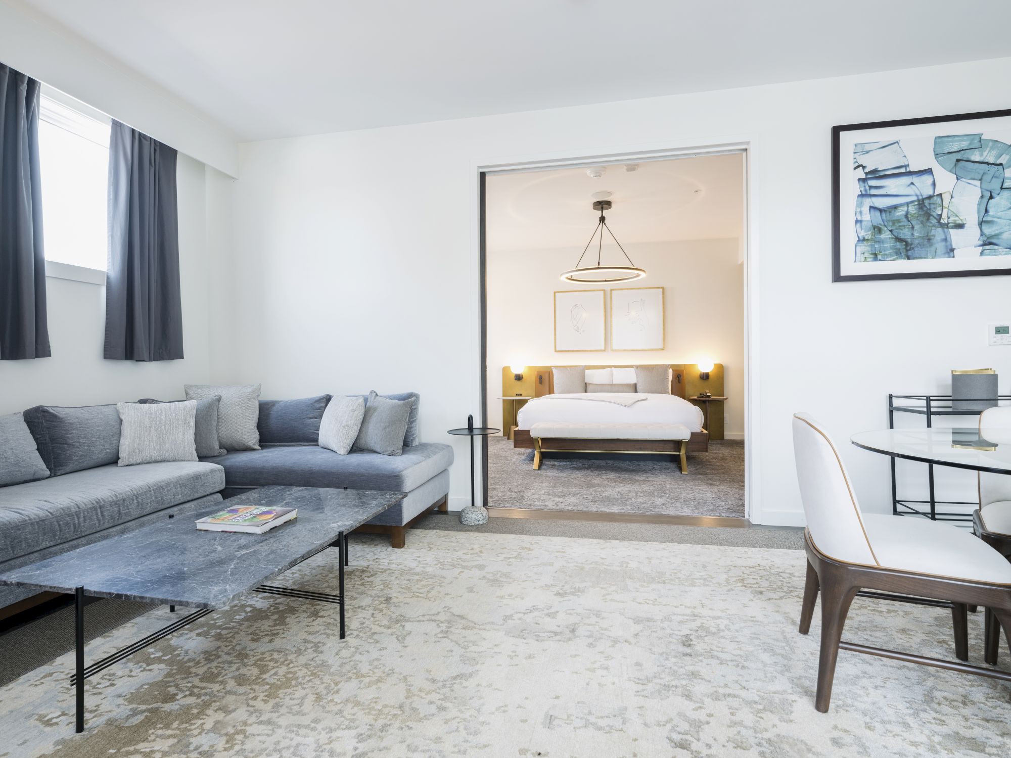 A modern living area with a gray couch, coffee table, and dining set opens into a bedroom with a neatly made bed and stylish decor.